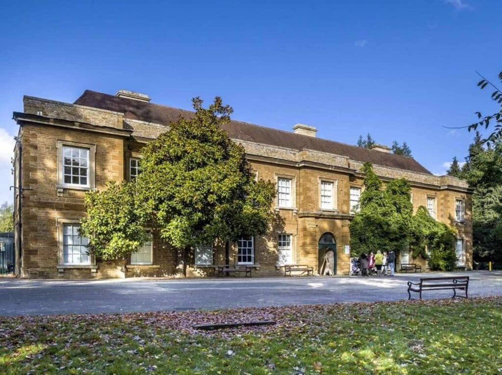colour photo of the exterior of Abington Park Museum building with grass and a bench on the right