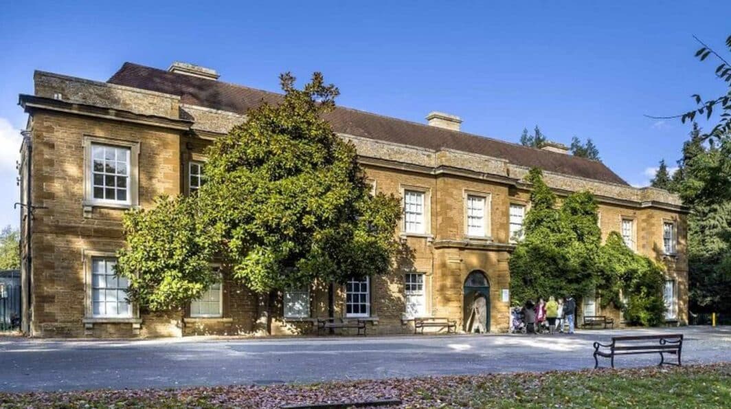 colour photo of the exterior of Abington Park Museum building with grass and a bench on the right