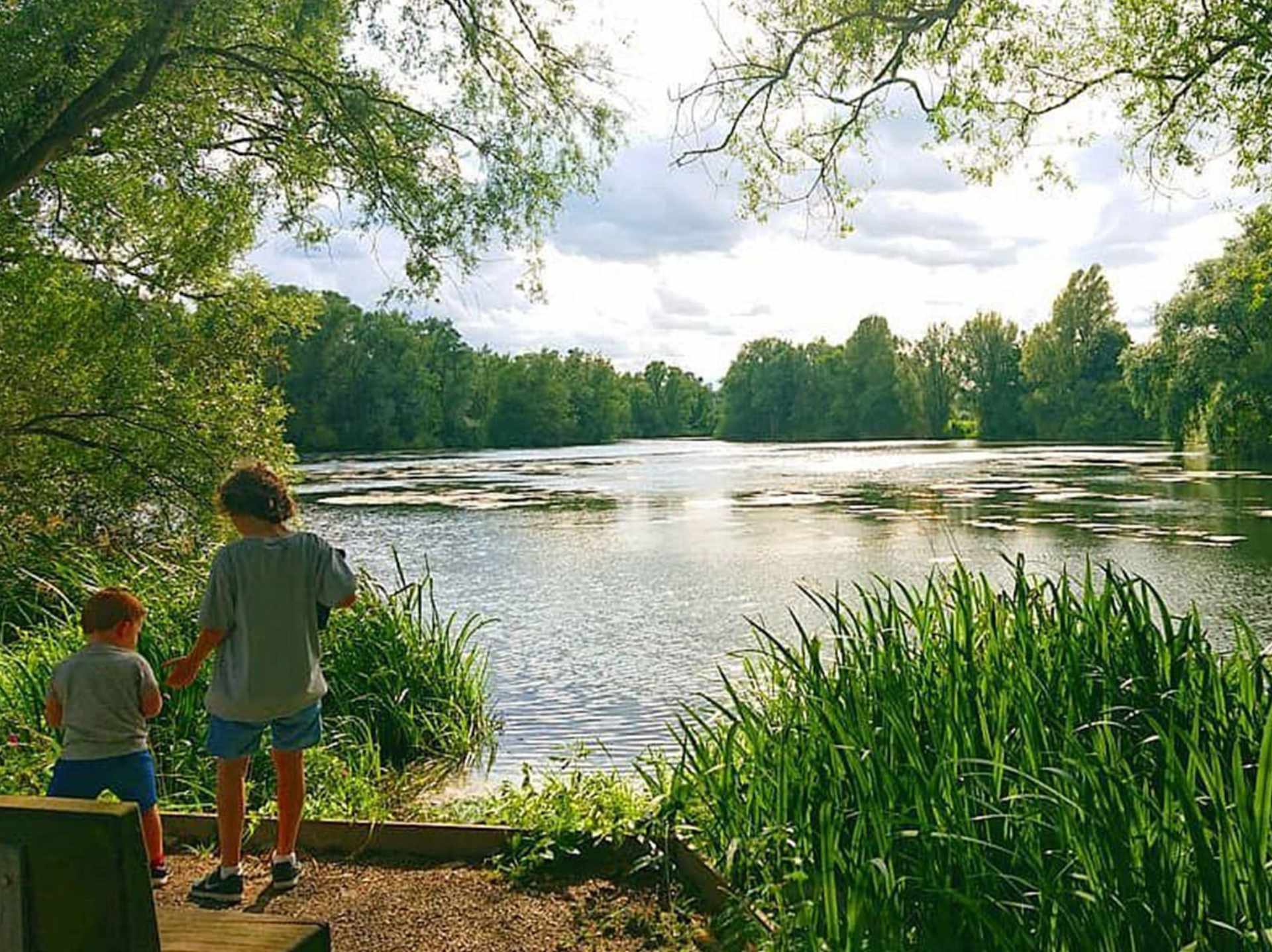 colour photo of two children standing in front of a lake with green trees and plants framing the image