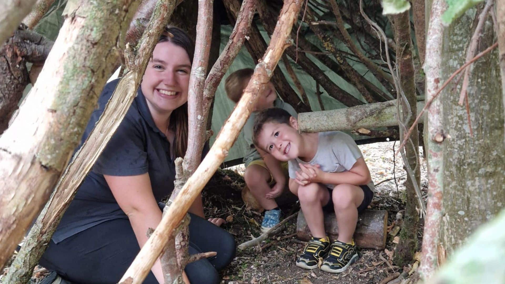 A ranger and two children hide in a den made out of sticks, all of them smiling.